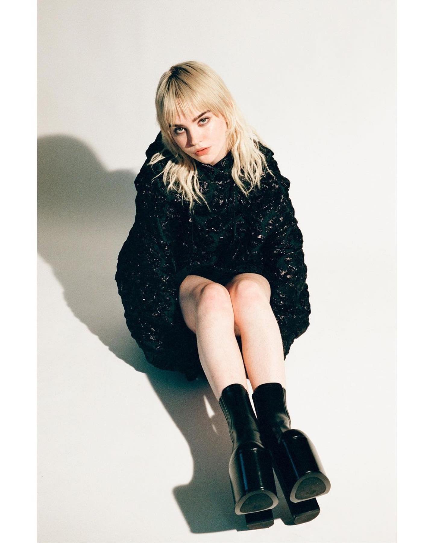 Young woman with blonde hair poses in dark, textured outfit and high-heeled boots against a white background in a studio portrait.