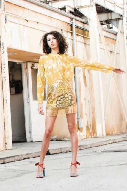 Woman poses in patterned yellow dress with floral details, red heels, standing outdoors against an industrial building backdrop.