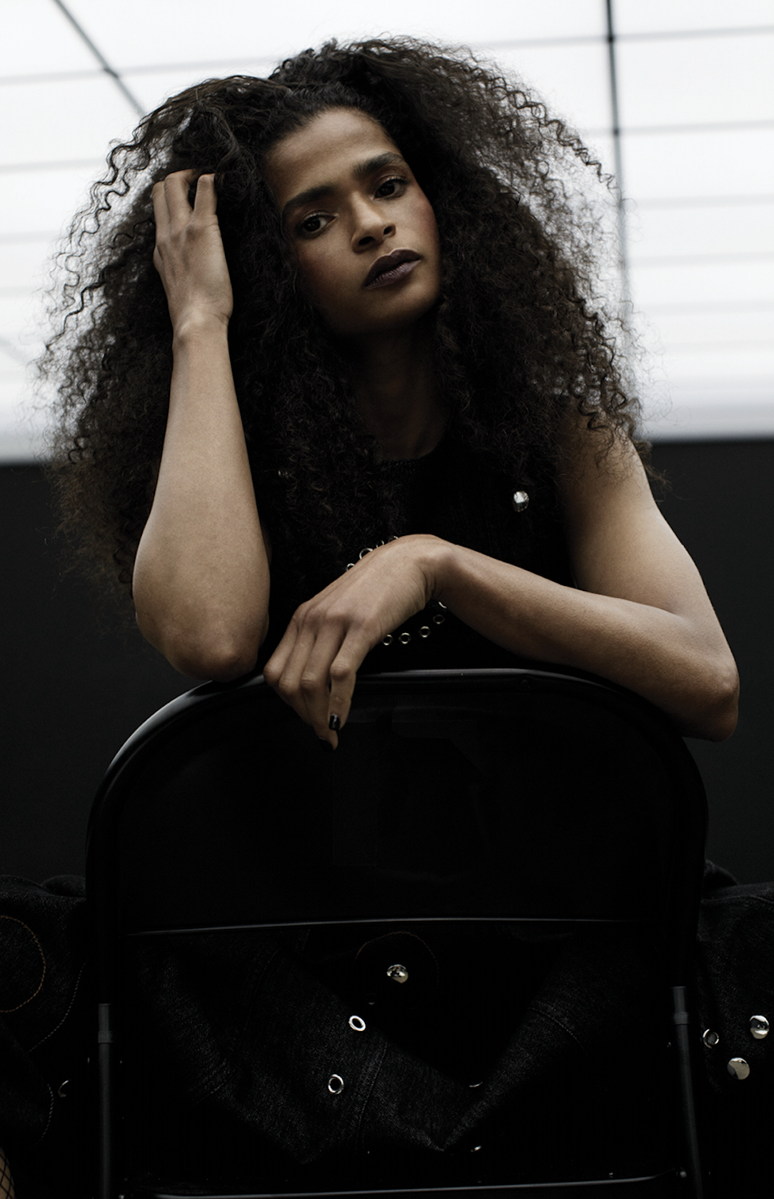 Adult female model with afro poses in front of black background, seated on black metal folding chair, looking at camera.