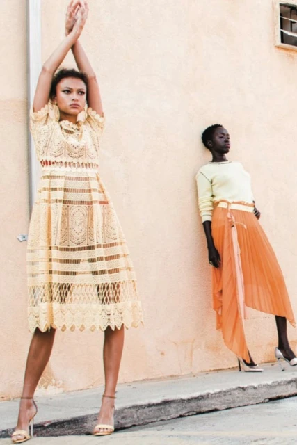 Two fashion models in dresses pose against a wall, one in a lacy ivory dress with hands raised, and the other in an orange skirt.