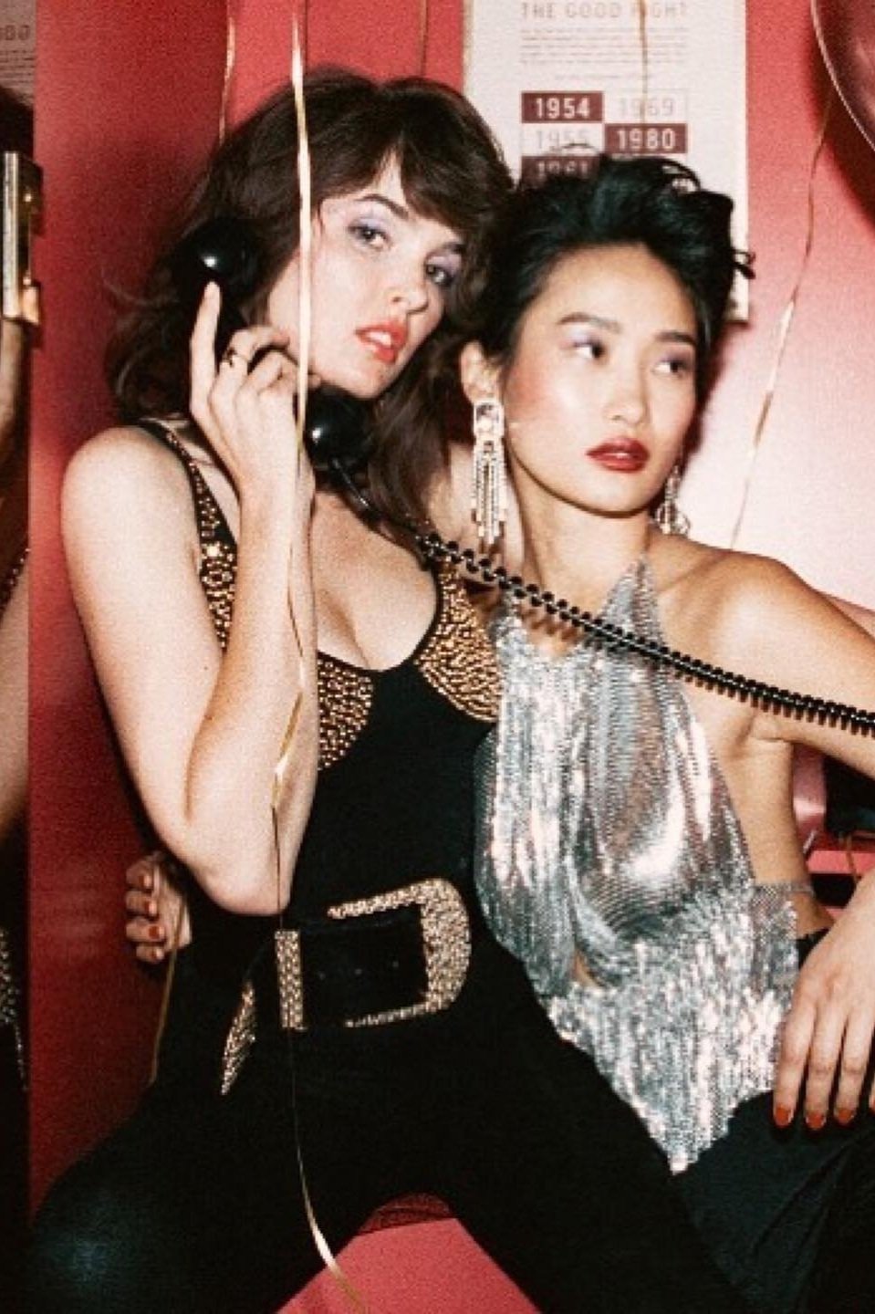 Two stylish young women in a photo booth, one talking on a vintage phone, wearing retro-inspired fashion and jewelry in warm, saturated light.