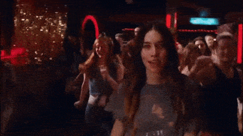 Crowd of adults dancing together indoors in a nightclub setting with red lighting and a blurry background atmosphere.
