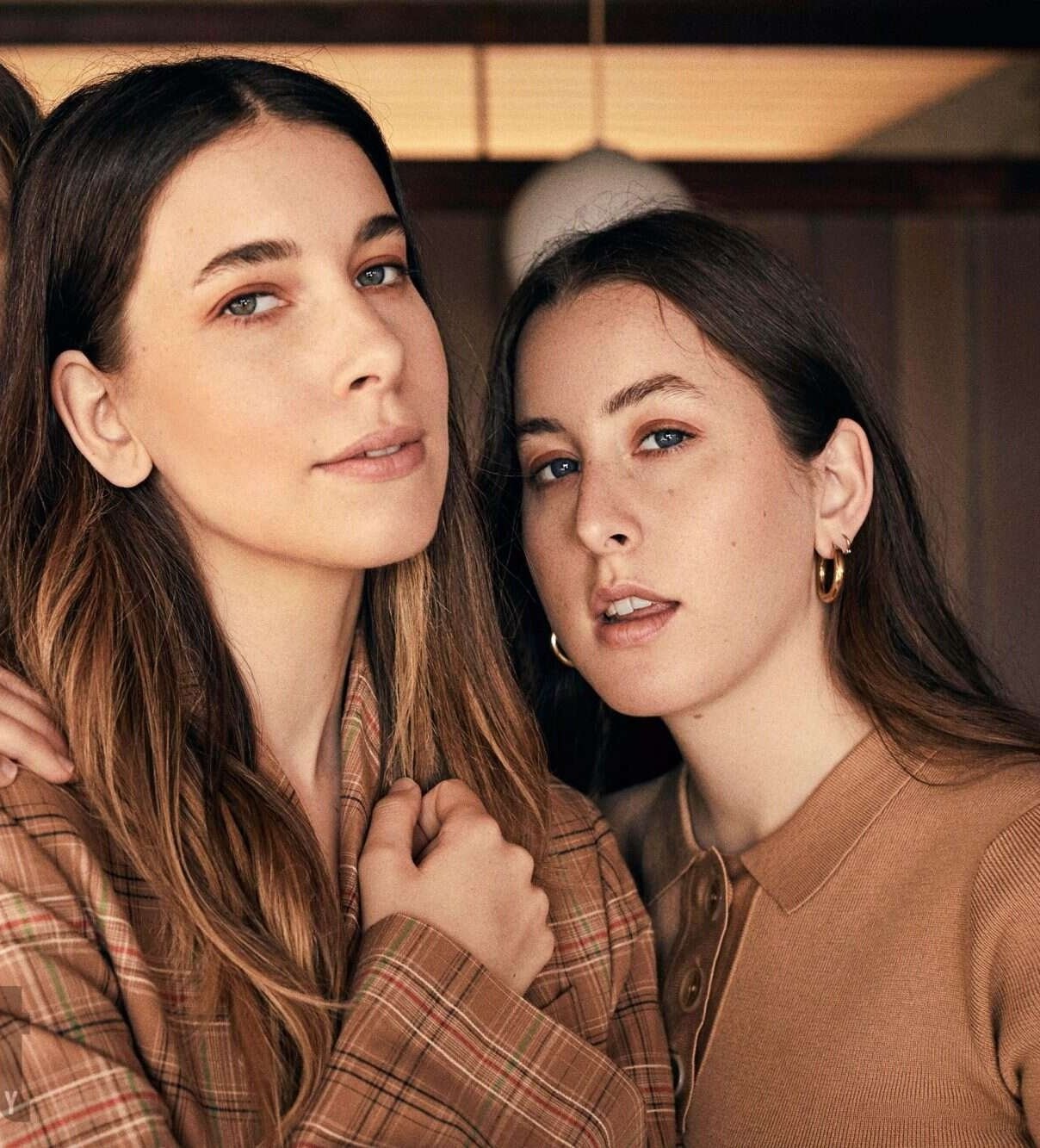 Two women pose indoors in a medium portrait shot. One wears a brown plaid jacket; the other wears a brown polo shirt. Both have long brown hair.