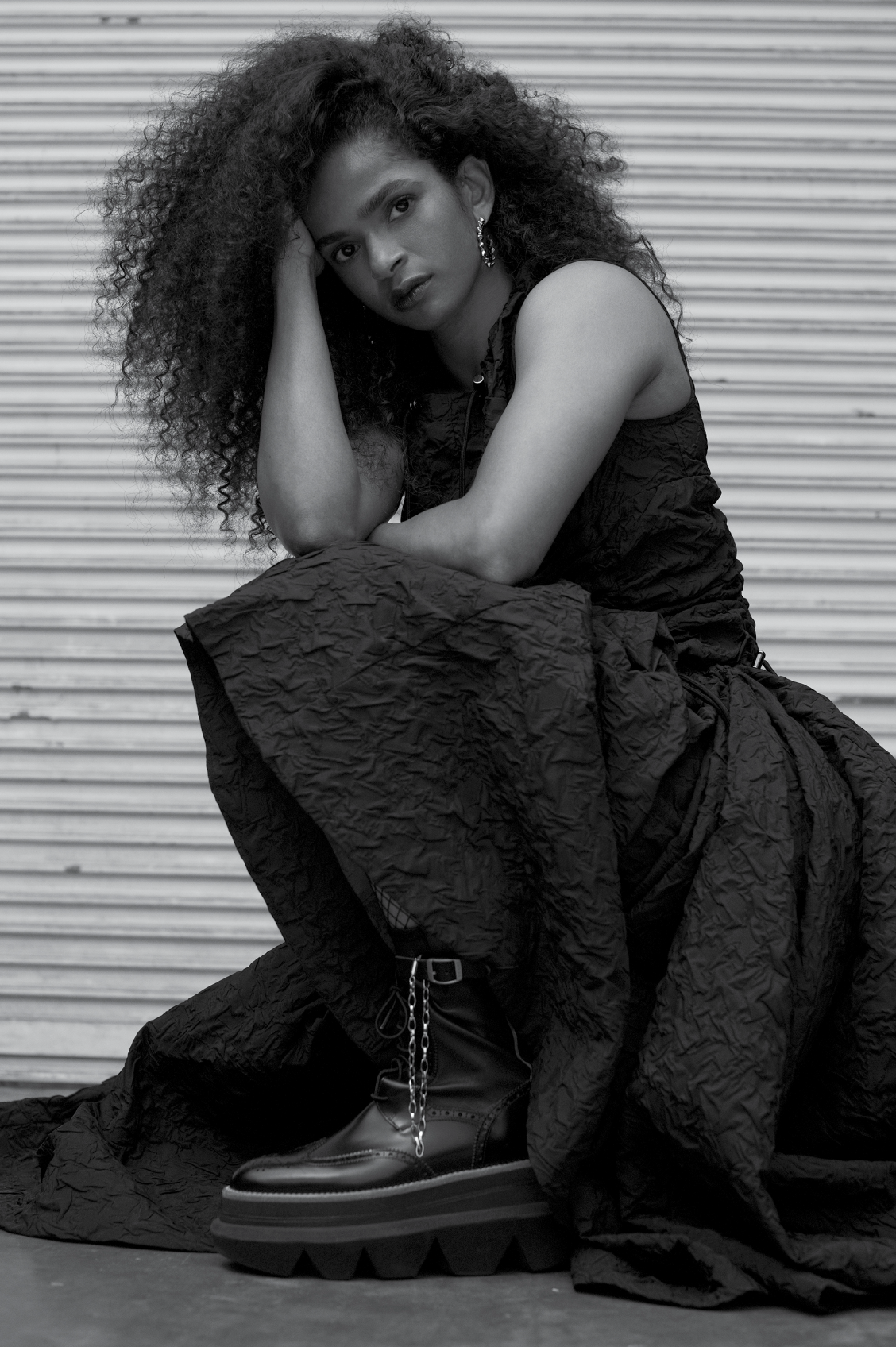 Woman with curly hair poses in black dress and platform boots against a roll-up metal door, looking directly at the camera.