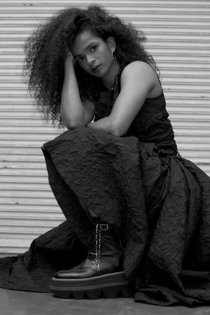 Woman with curly hair poses in black dress and platform boots against a roll-up metal door, looking directly at the camera.