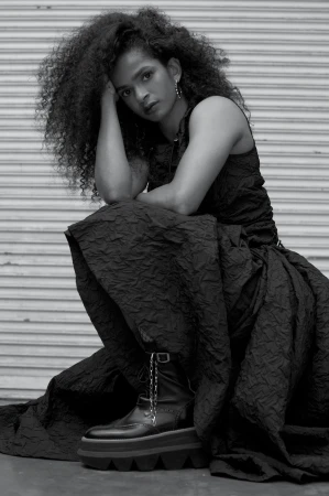 Woman with curly hair poses in black dress and platform boots against a roll-up metal door, looking directly at the camera.
