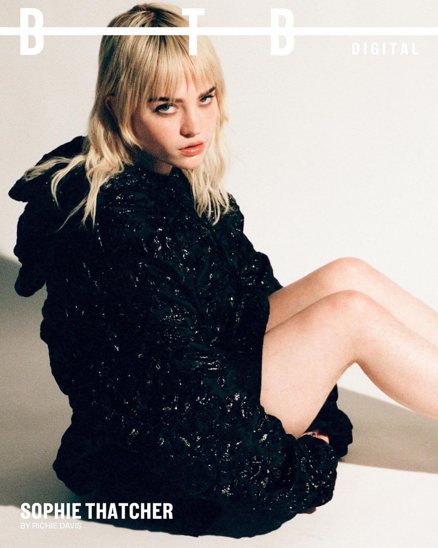 Blonde woman with bangs, wearing a black hooded jacket, poses in a studio against a white background for a fashion magazine cover.