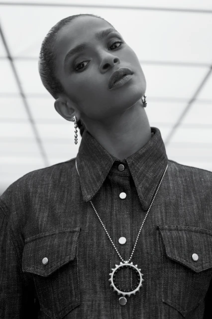 Black and white portrait of an African American woman in a denim shirt and silver necklace, posing against a white geometric background.
