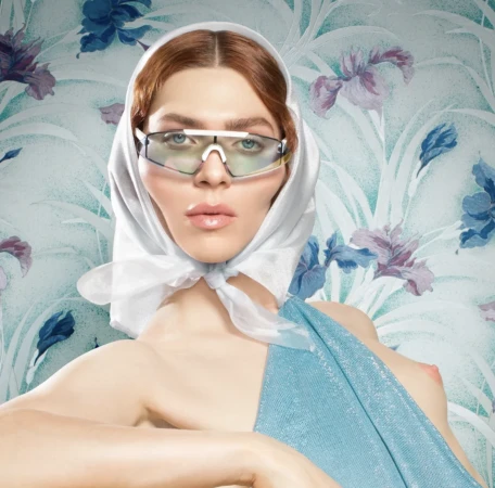 Model with red hair wearing futuristic wraparound sunglasses, a white headscarf, and a blue top against a floral wallpaper background.