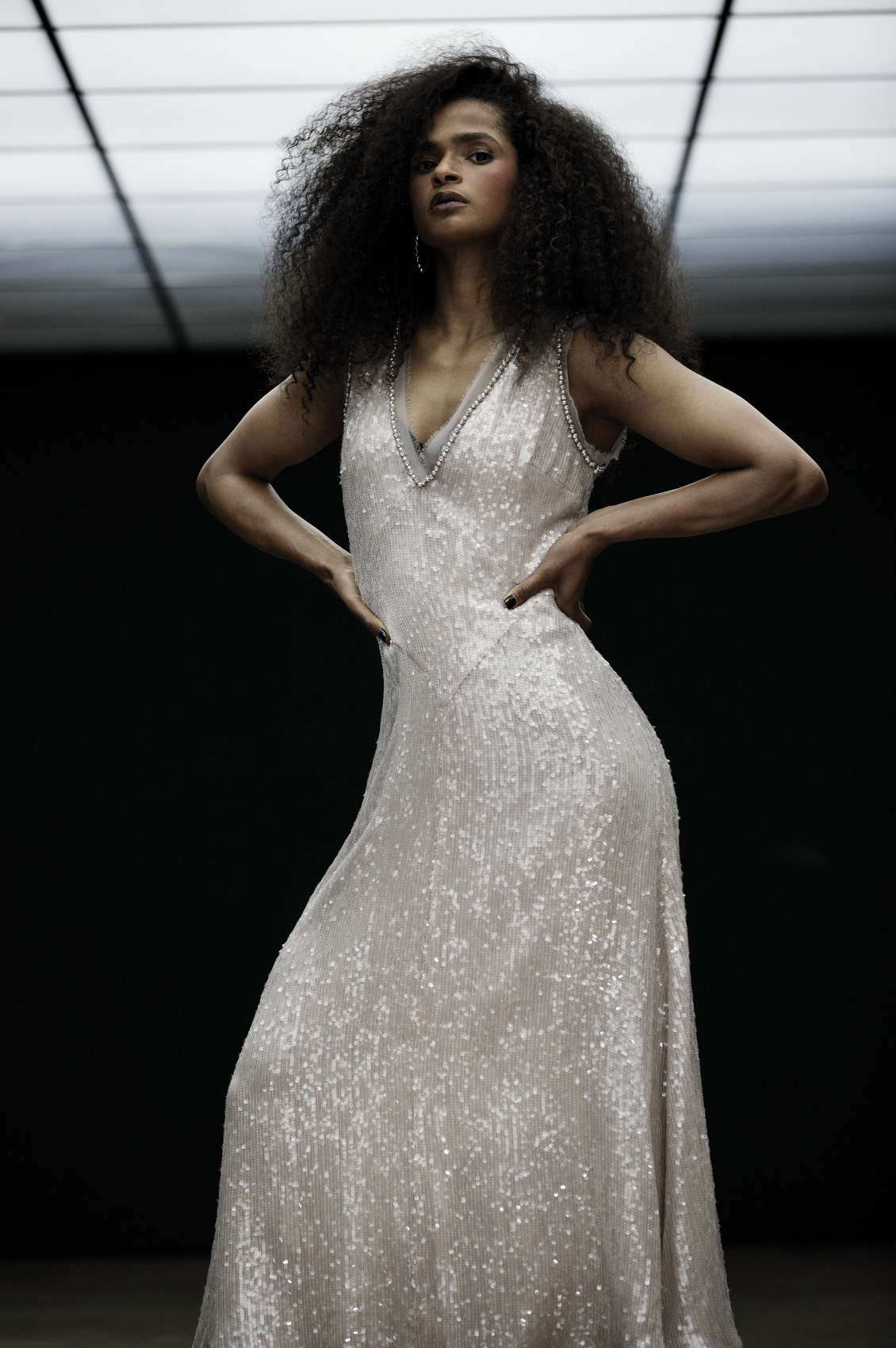 A glamorous woman with curly hair wears a long, shimmering gown with a V-neck in a dimly lit space with a glowing ceiling.