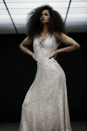 A glamorous woman with curly hair wears a long, shimmering gown with a V-neck in a dimly lit space with a glowing ceiling.