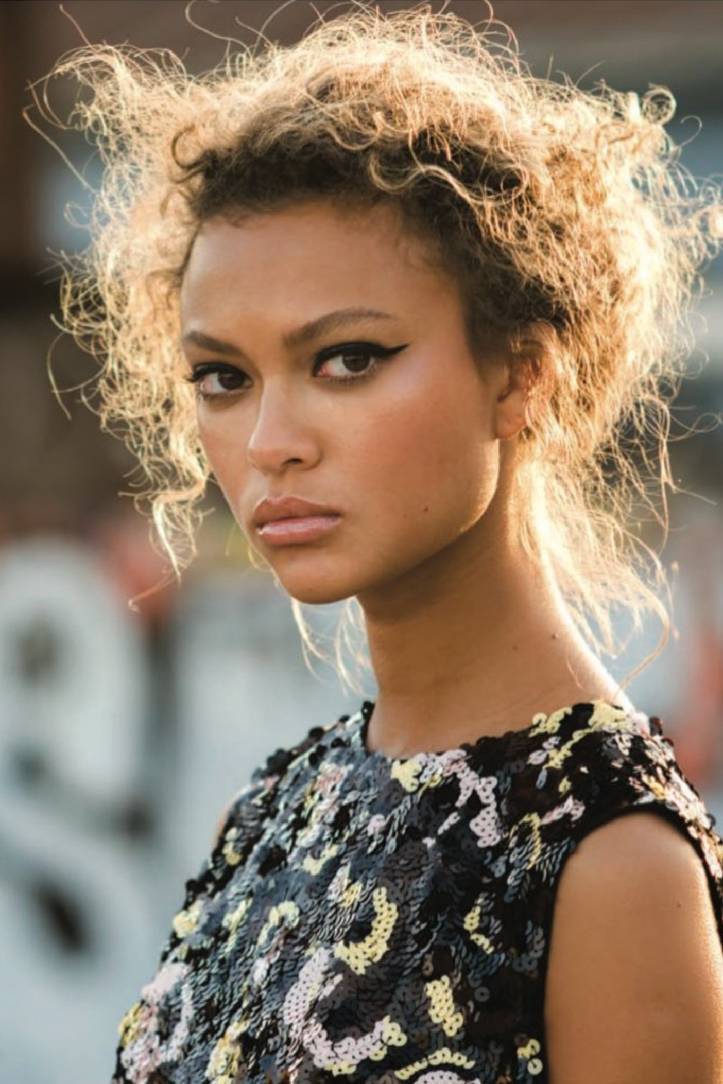 Close-up portrait of a stylish adult female model with curly blonde hair wearing a black sequined top against a blurred backdrop.