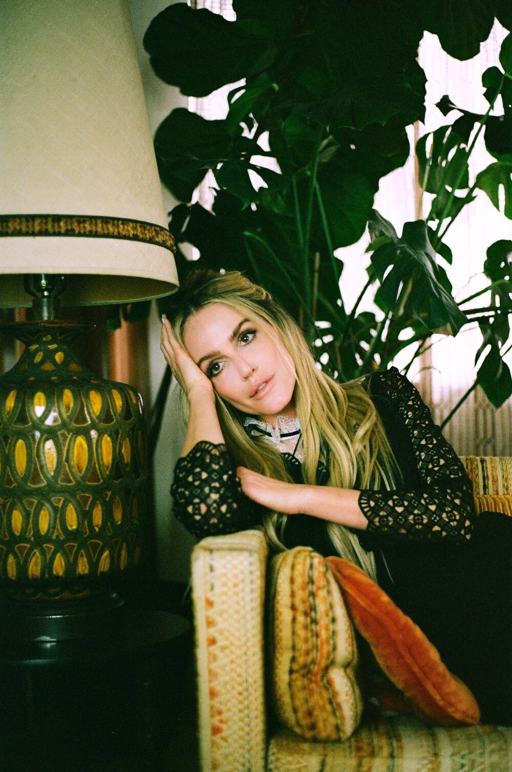 Blonde woman seated on a patterned sofa next to a large floor lamp and indoor house plants