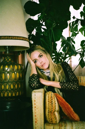 Blonde woman seated on a patterned sofa next to a large floor lamp and indoor house plants