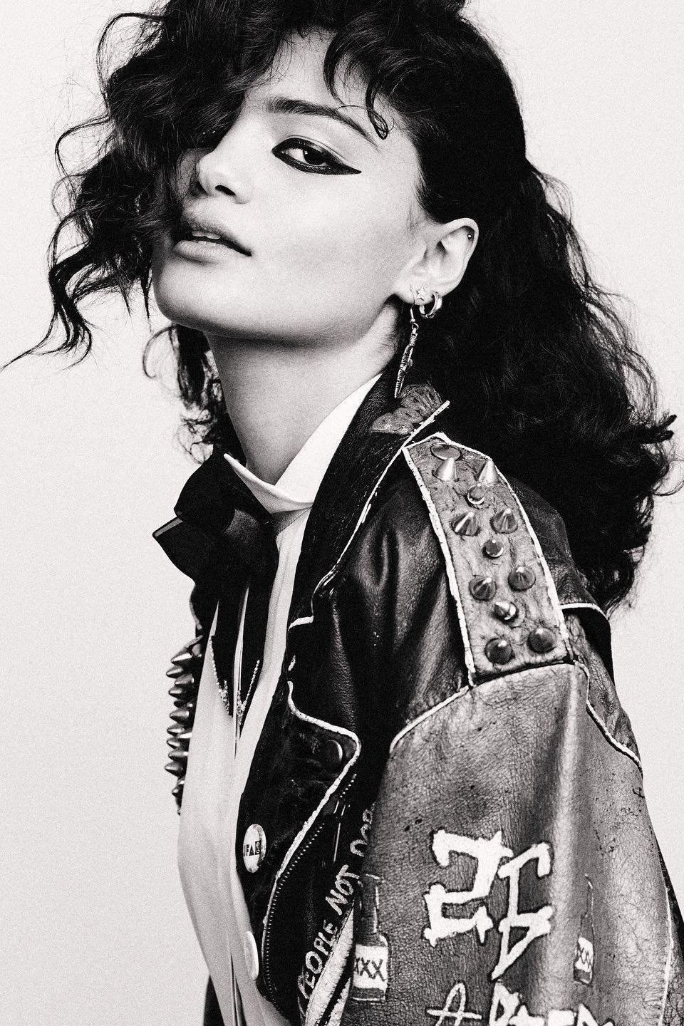 Monochrome portrait of a young woman with dark curly hair, dramatic eyeliner, and a studded leather jacket in a light grey setting.