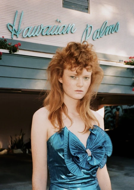 A red-haired woman in a blue dress stands in front of the Hawaiian Palms building, with a close-up shot capturing her pose and facial expression.