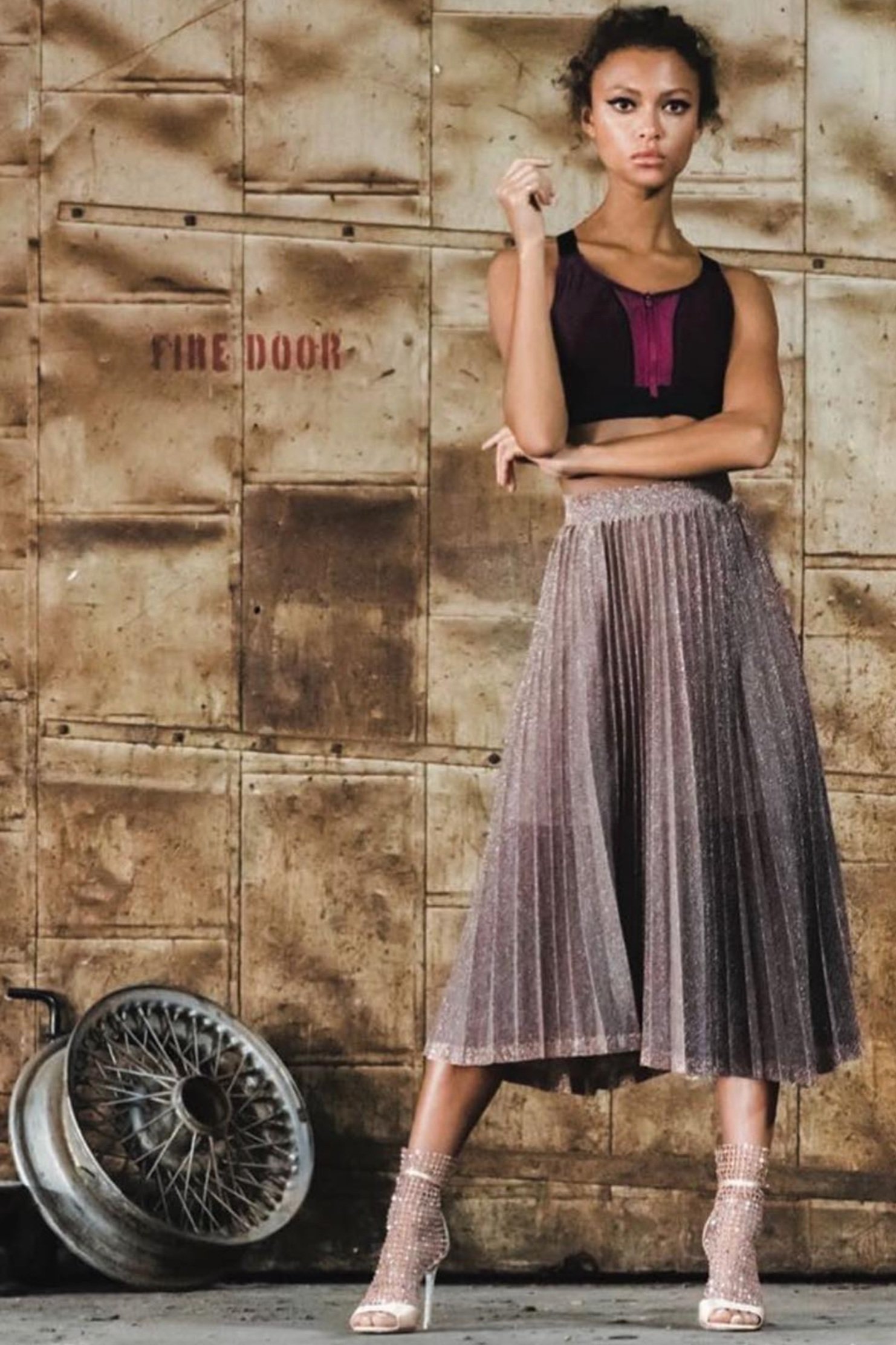 A young adult woman poses in a crop top and midi skirt, standing near a tire in front of a weathered industrial door with a fire door sign.