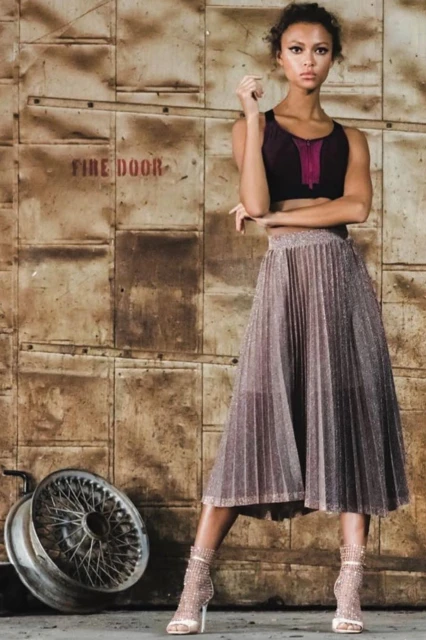 A young adult woman poses in a crop top and midi skirt, standing near a tire in front of a weathered industrial door with a fire door sign.