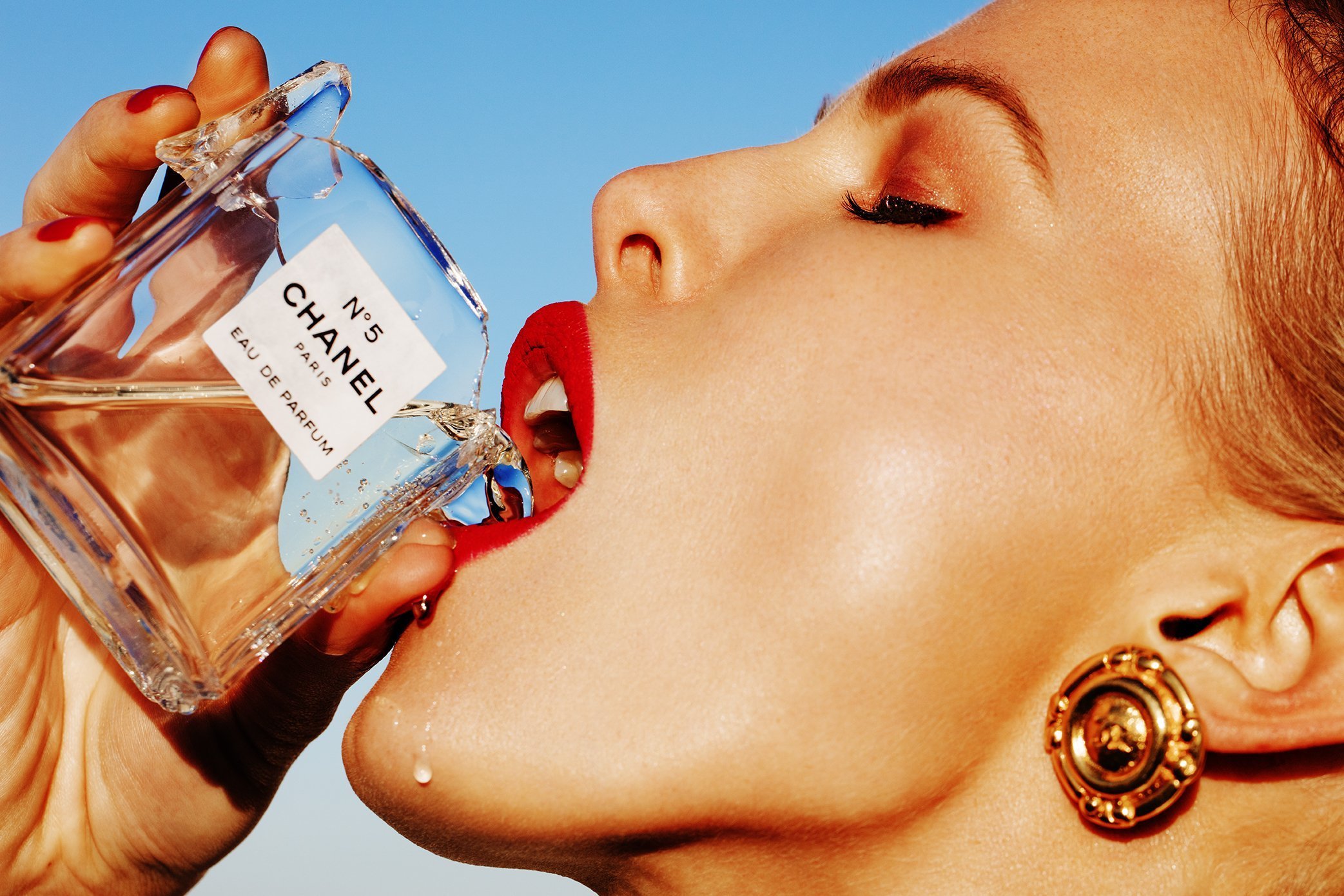 Close-up of a fair-skinned woman with red lipstick drinking Chanel N°5 perfume, against a bright blue sky backdrop.