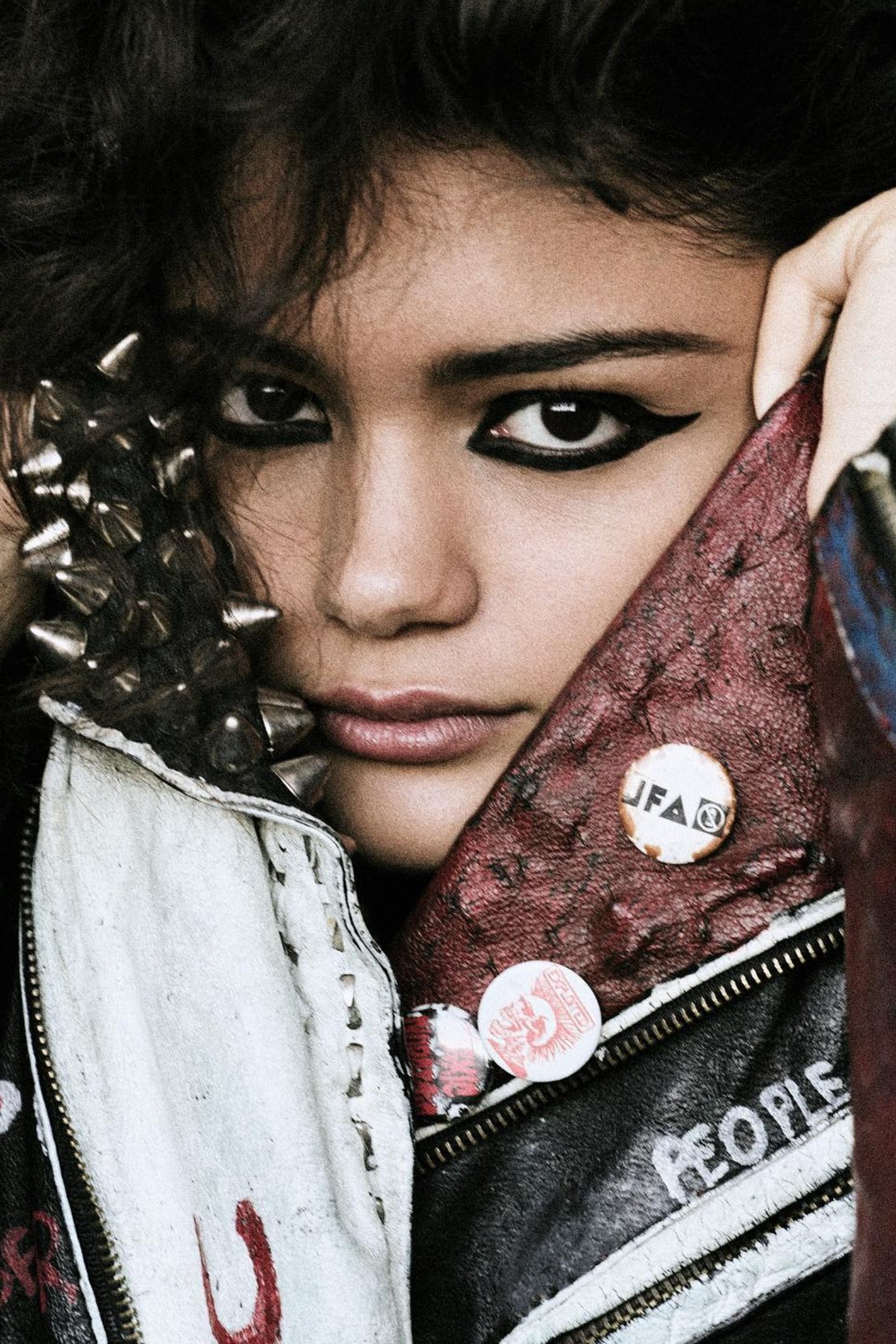 Close-up portrait of a young adult woman with dark hair, striking eye makeup, and a studded leather jacket with pins.