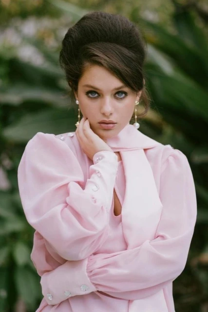 Portrait of a woman with a large bouffant hairstyle, wearing a pale pink vintage-style dress with pearl earrings against a green backdrop.