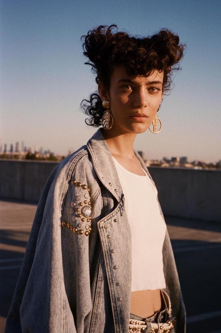 Woman with curly updo poses in sunlight wearing a white top and embellished denim jacket against a city skyline background.