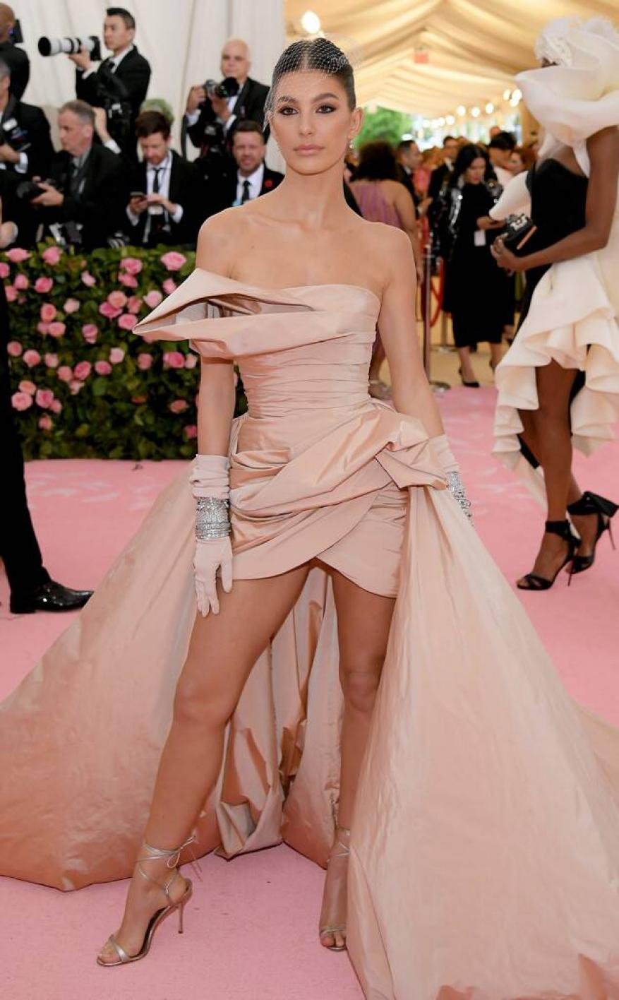 Woman on pink carpet in blush strapless gown with long train, strappy heels, and elbow-length gloves poses at a red carpet event.