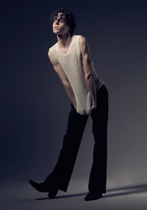 Young man with curly dark hair wearing a ribbed cream knit tank top and black trousers against a grey studio background