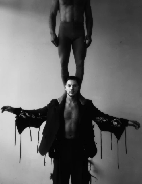 Black and white image of a shirtless man with dark hair holding up another shirtless man in red bottoms on his shoulders.