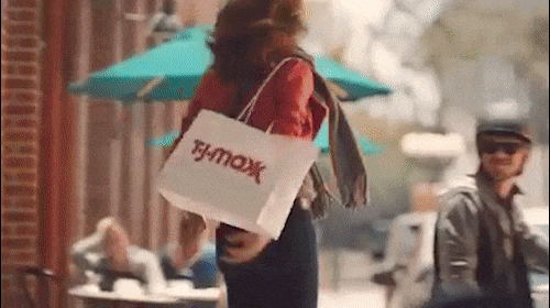 Adult female walking with TJ Maxx shopping bag on a city street with other people and buildings in the background on a sunny day.