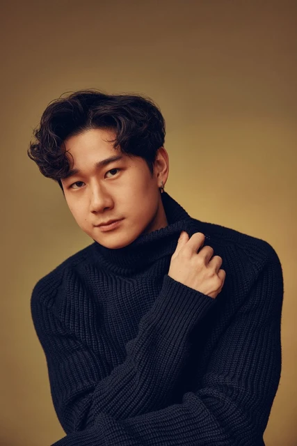 Portrait of an Asian man with dark curly hair, wearing a navy blue turtleneck sweater, posing against a neutral golden background.