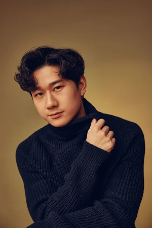 Portrait of an Asian man with dark curly hair, wearing a navy blue turtleneck sweater, posing against a neutral golden background.