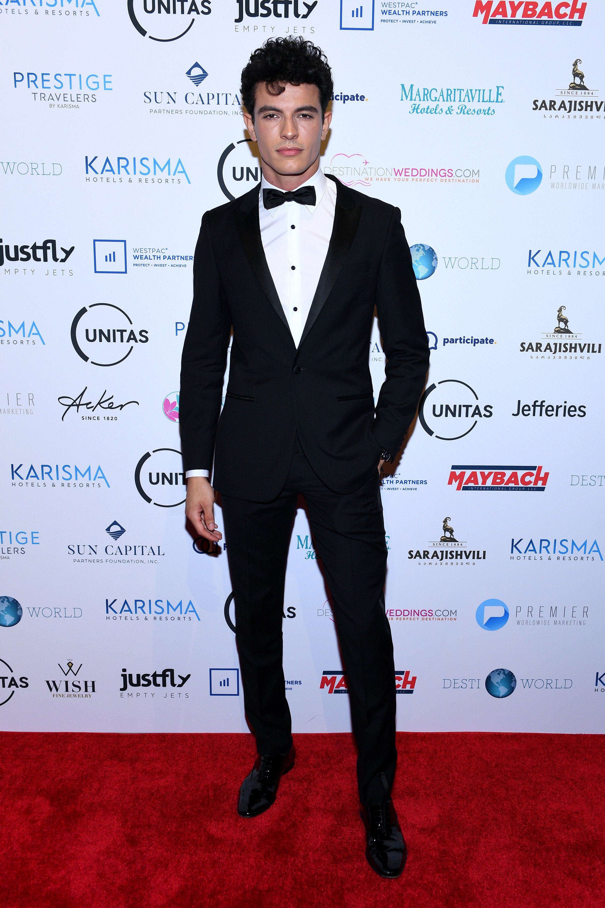 Man in a black suit and bow tie poses on a red carpet in front of logos for Karisma, Margaritaville, Maybach, and other sponsors.