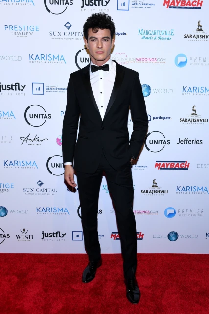 Man in a black suit and bow tie poses on a red carpet in front of logos for Karisma, Margaritaville, Maybach, and other sponsors.