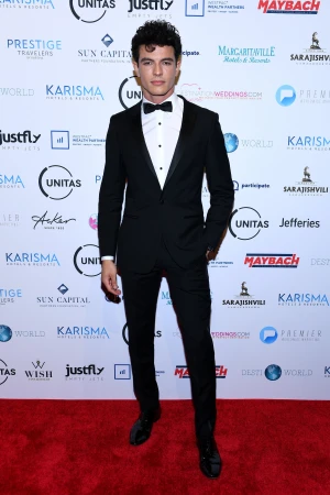 Man in a black suit and bow tie poses on a red carpet in front of logos for Karisma, Margaritaville, Maybach, and other sponsors.