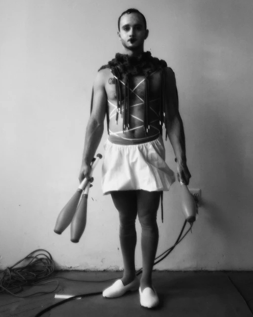 Monochrome portrait of a bare-chested performer with face paint, wearing a skirt, feather boa, and holding juggling clubs indoors.