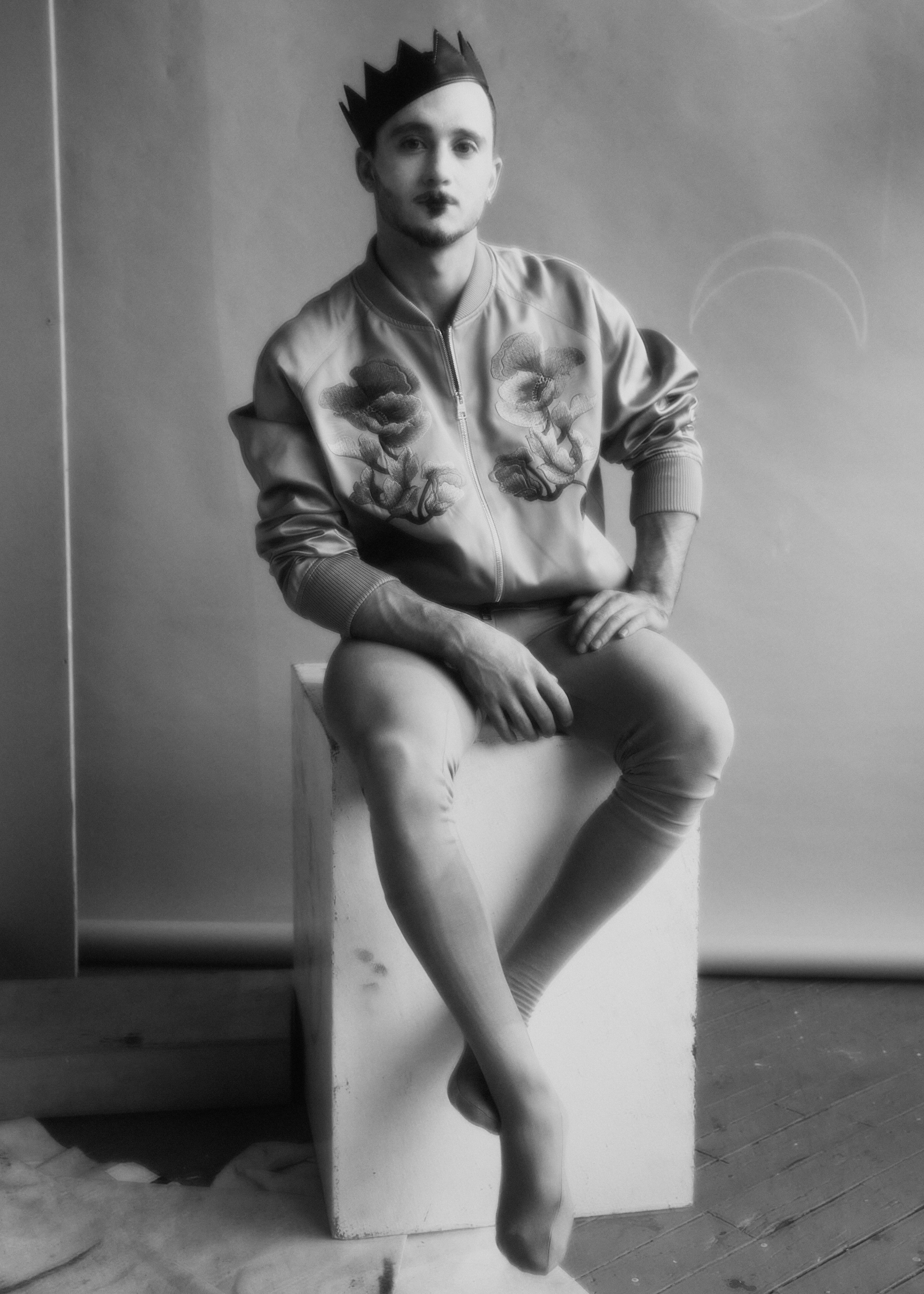 Monochrome portrait of a male figure with painted face, wearing a crown and suit, seated on a white box against a grey backdrop.