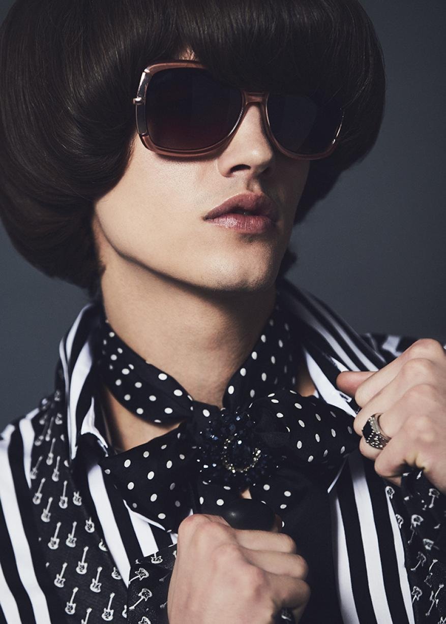 Close-up portrait of a model with dark bowl cut hair, oversized sunglasses, striped shirt and polka dot necktie.
