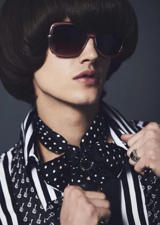 Close-up portrait of a model with dark bowl cut hair, oversized sunglasses, striped shirt and polka dot necktie.