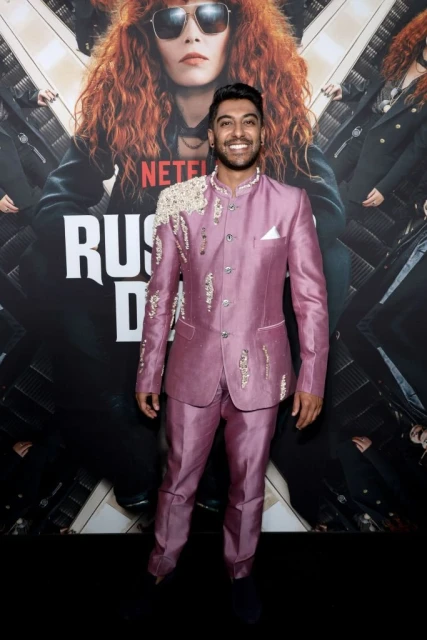 Man in a mauve suit poses in front of a Russian Doll promotional poster featuring a woman with red hair and sunglasses, smiling.
