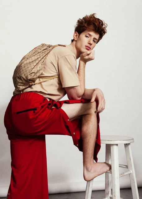 Red-haired male fashion model with beige t-shirt and red pants posing barefoot on white stool in front of white background in studio.