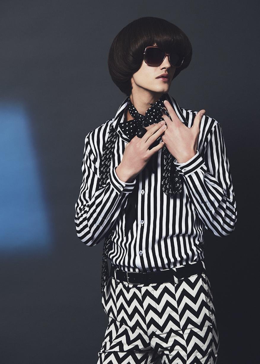 A young adult with an elegant hairstyle wears sunglasses, a striped shirt, and patterned pants against a dark blue gradient backdrop.