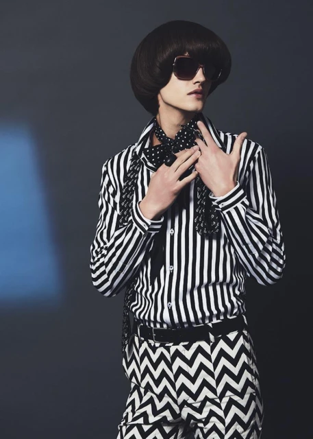 A young adult with an elegant hairstyle wears sunglasses, a striped shirt, and patterned pants against a dark blue gradient backdrop.