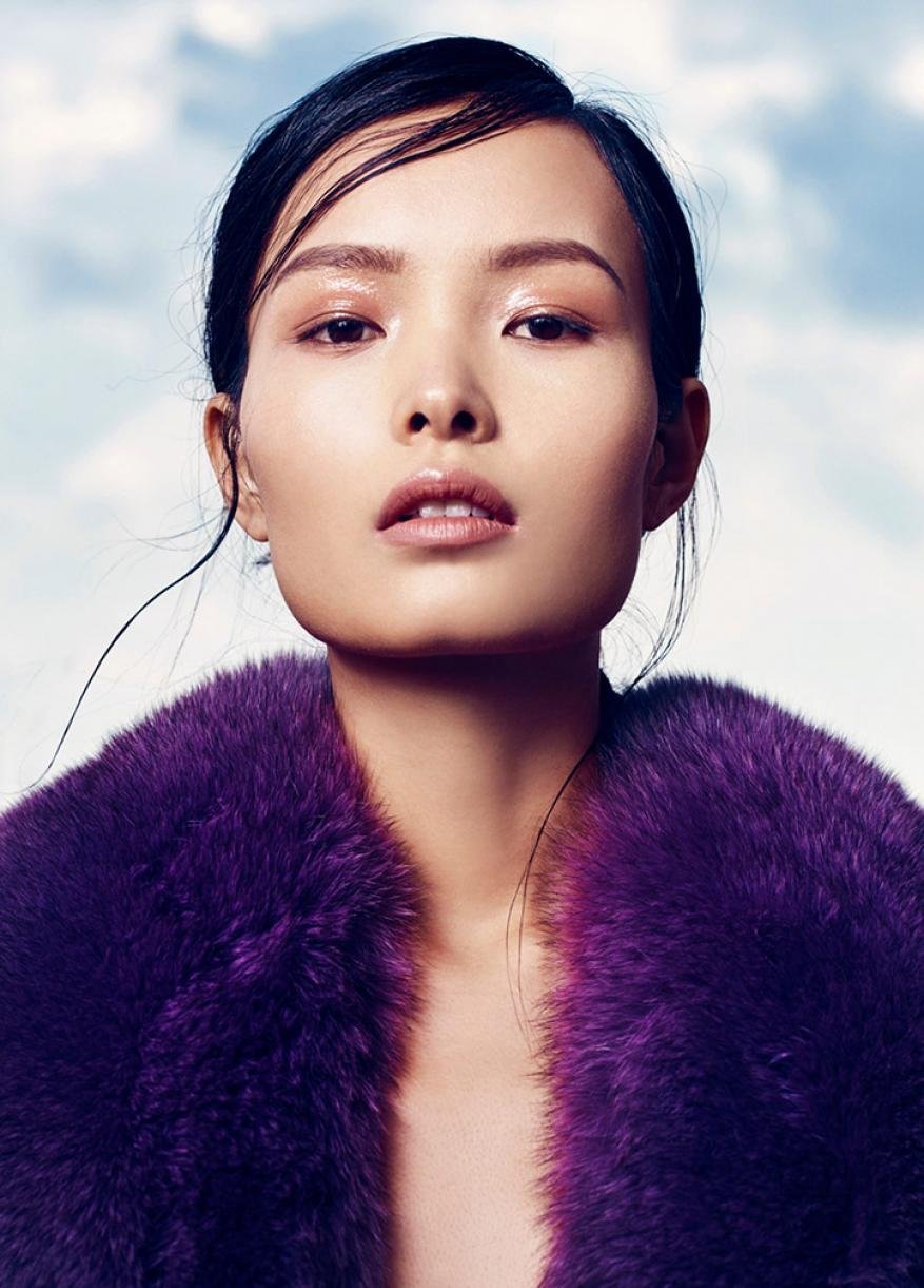 Glamorous Asian woman with flawless skin and a purple fur coat poses against a backdrop of clouds. Beauty and fashion portrait.