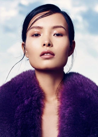 Glamorous Asian woman with flawless skin and a purple fur coat poses against a backdrop of clouds. Beauty and fashion portrait.