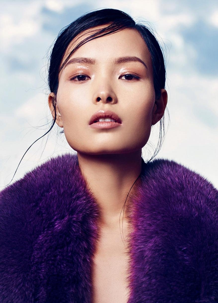 Close-up fashion shot of an Asian female model wearing a vibrant purple faux fur coat against a soft, diffused blue and white sky background.