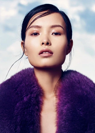 Close-up fashion shot of an Asian female model wearing a vibrant purple faux fur coat against a soft, diffused blue and white sky background.