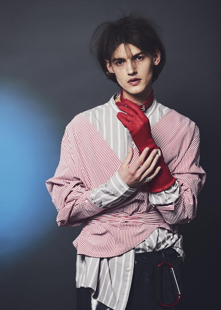 Fashion portrait of a young adult wearing a striped shirt and gloves, posing in a studio with soft lighting and a modern aesthetic.