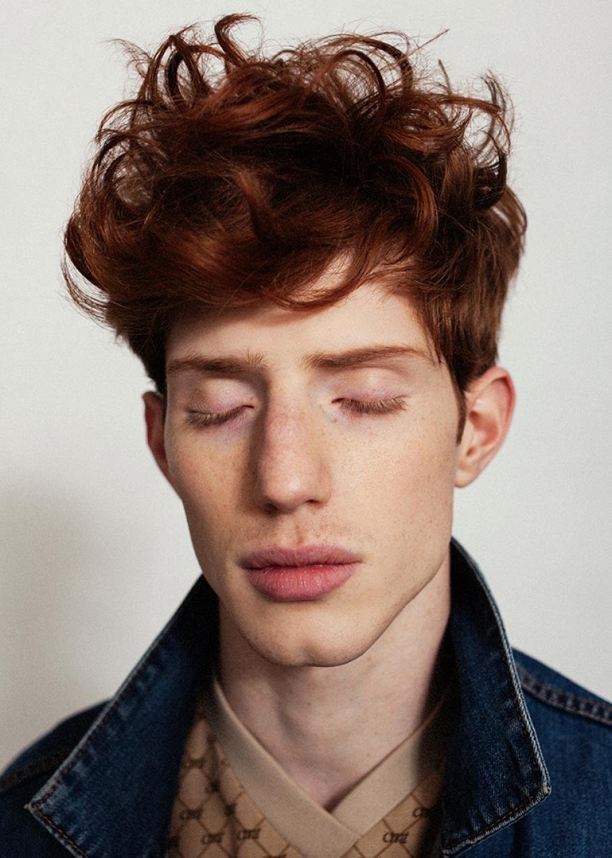 Portrait of a man with curly red hair and closed eyes wearing a denim jacket and patterned shirt against a white background