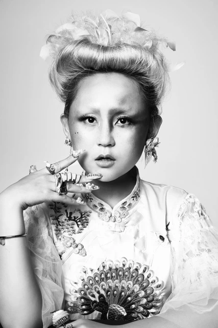 Black and white portrait of an Asian woman with elaborate makeup, hairstyle, and jewelry against a light background in a studio.